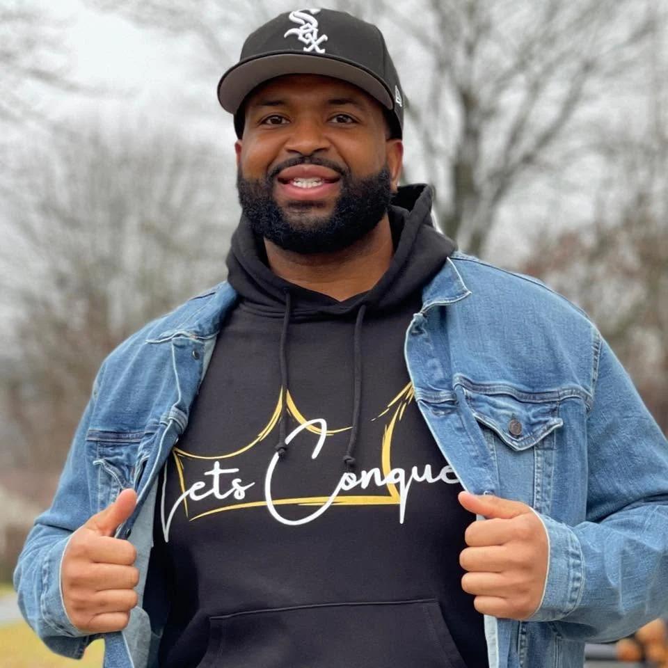 Outdoor portrait of a person wearing a denim jacket, dark hoodie, and baseball cap